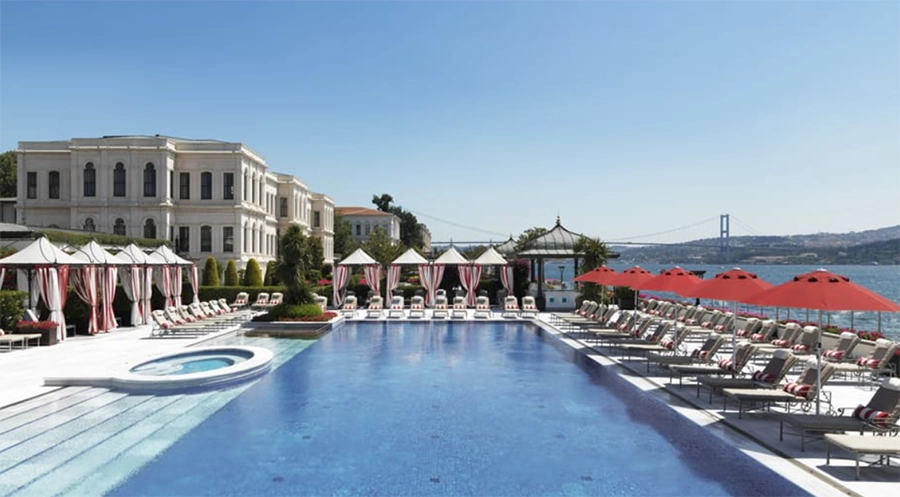 Four Seasons Bosphorus otelinin Boğaz manzaralı açık havuzunda, kırmızı-beyaz çizgili tenteler ve şık şezlonglar serin bir yaz kaçamağı için beklerken, arka planda zarif bir köşk ve Boğaz Köprüsü'nün silüeti İstanbul'un en iyi otel havuzlarından birini gözler önüne seriyor.