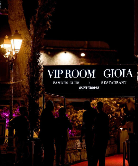 Saint-Tropez'de hareketli bir geceyi yansıtan bu fotoğrafta, VIP Room adlı ünlü kulüp ve Gioia restoranının girişinde kırmızı halı üzerinde silüetler halinde toplanmış insanlar görülüyor; loş sokak lambalarının ve mor-turuncu ambiyans ışıklarının aydınlattığı bu mekan, Fransız Rivierası'nın lüks ve canlı eğlence atmosferini sergiliyor.