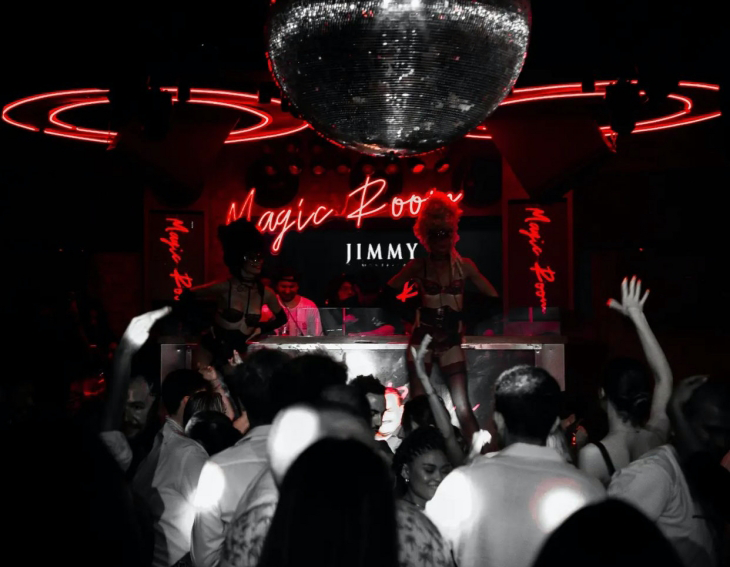 Kırmızı neon ışıkları ve tavandan sarkan büyük disko topuyla aydınlatılmış, Magic Room ve Jimmy tabelalarının da göründüğü loş bir gece kulübünde, sahnedeki iki performans sanatçısı ve bir DJ eşliğinde coşkuyla dans eden kalabalık bir grup insan tasvir ediliyor.