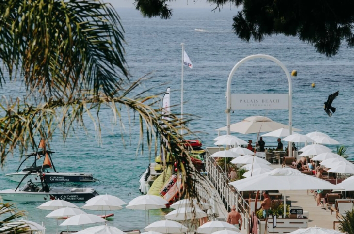 Palmiye ağaçları ve masmavi gökyüzü altında, Fransız Rivierası'nın kristal sularında yer alan hareketli Palais Beach; beyaz şemsiyelerle dolu bir iskelede ve kıyıda güneşlenen insanlar, yüzen tekneler ve tatil atmosferiyle göz alıcı bir yaz manzarası sunuyor.