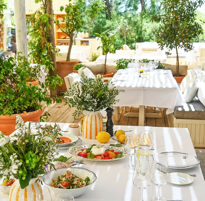 Güneşli bir dış mekan restoranında, beyaz örtülü masalar taze salatalar, burrata peyniri ve domateslerle dolu tabaklar gibi lezzetli başlangıçlarla donatılmış, turuncu çizgili vazolar, limonlar ve taze yeşil bitkilerle çevrili davetkar bir yemek deneyimi sunuyor.