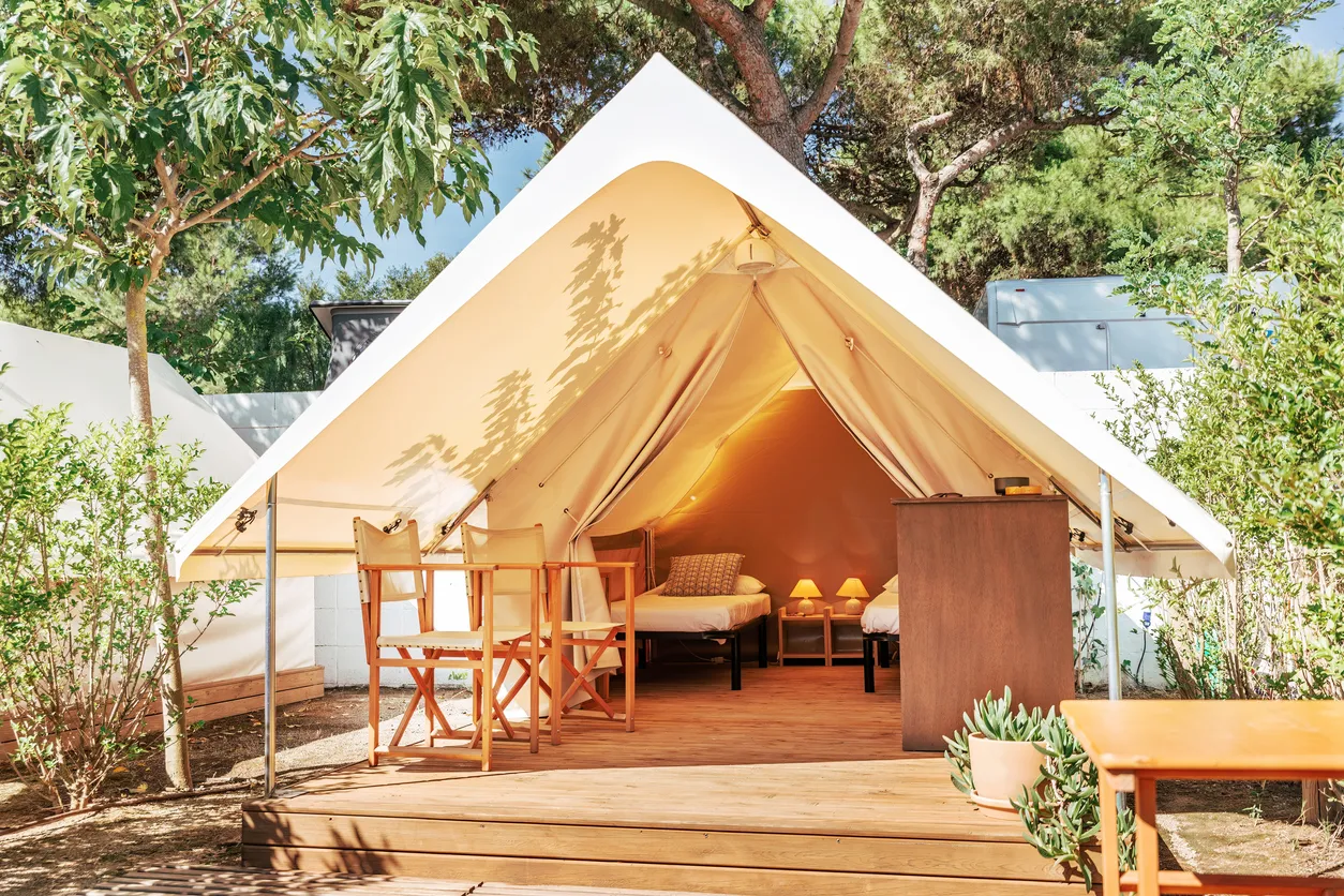 Aydınlık bir havada, yemyeşil ağaçlar ve bitkilerle çevrili bir alanda, önü açık, ahşap bir veranda üzerine kurulmuş büyük, bej renkli bir glamping çadırı, içerisinde iki tek kişilik yatak, komodinler ve sıcak ışık veren lambalarla döşenmiş konforlu iç mekanı ve önündeki katlanır sandalyelerle lüks bir doğa konaklaması sunuyor.