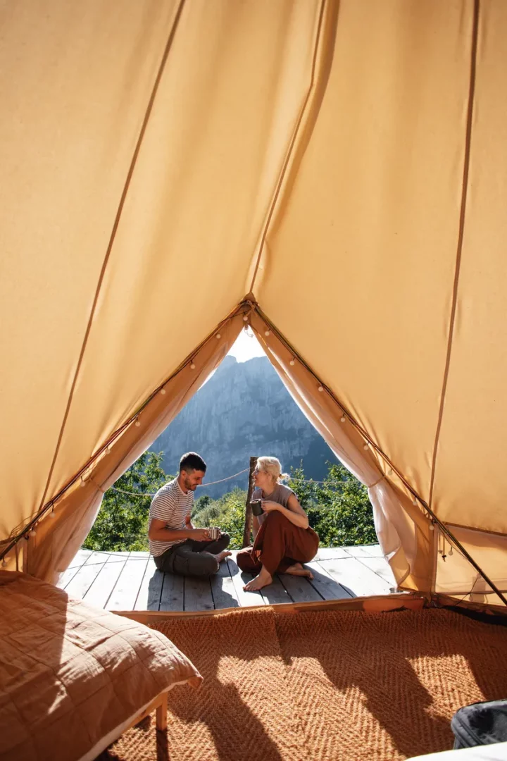 Bej renkli ferah bir glamping çadırının içinden çekilen bu karede, ahşap zeminde karşılıklı oturan ve dağ manzarasına karşı kahvelerini yudumlayan bir çift, yeşillikler ve yükselen dağlarla çevrili huzurlu bir doğa tatili anının keyfini çıkarıyor.
