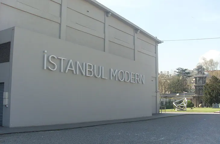 Gri dış cephede büyük harflerle “İSTANBUL MODERN” yazan tabelanın ve müze giriş alanının önü açık bir avlu ile görünümü.