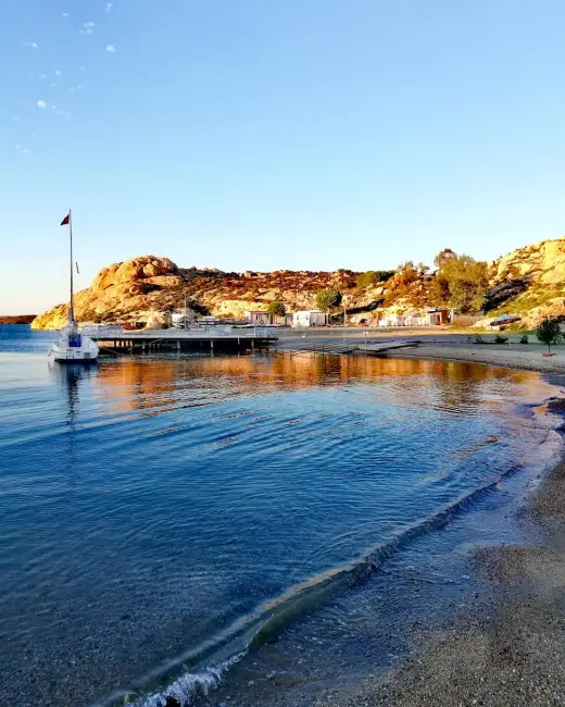 Ege'nin sevilen rotası Foça'daki Karakum Plajı'nda, gün batımının kayalık yamaçlara vuran altın rengi ışıklarıyla parlayan, temiz mavi denizin hafifçe kıyıya vuran dalgaları, kum ve çakıl karışımı sahili, iskelede demirli beyaz bir yelkenli ve kıyıdaki tesislerle huzurlu bir akşamüstü manzarası.