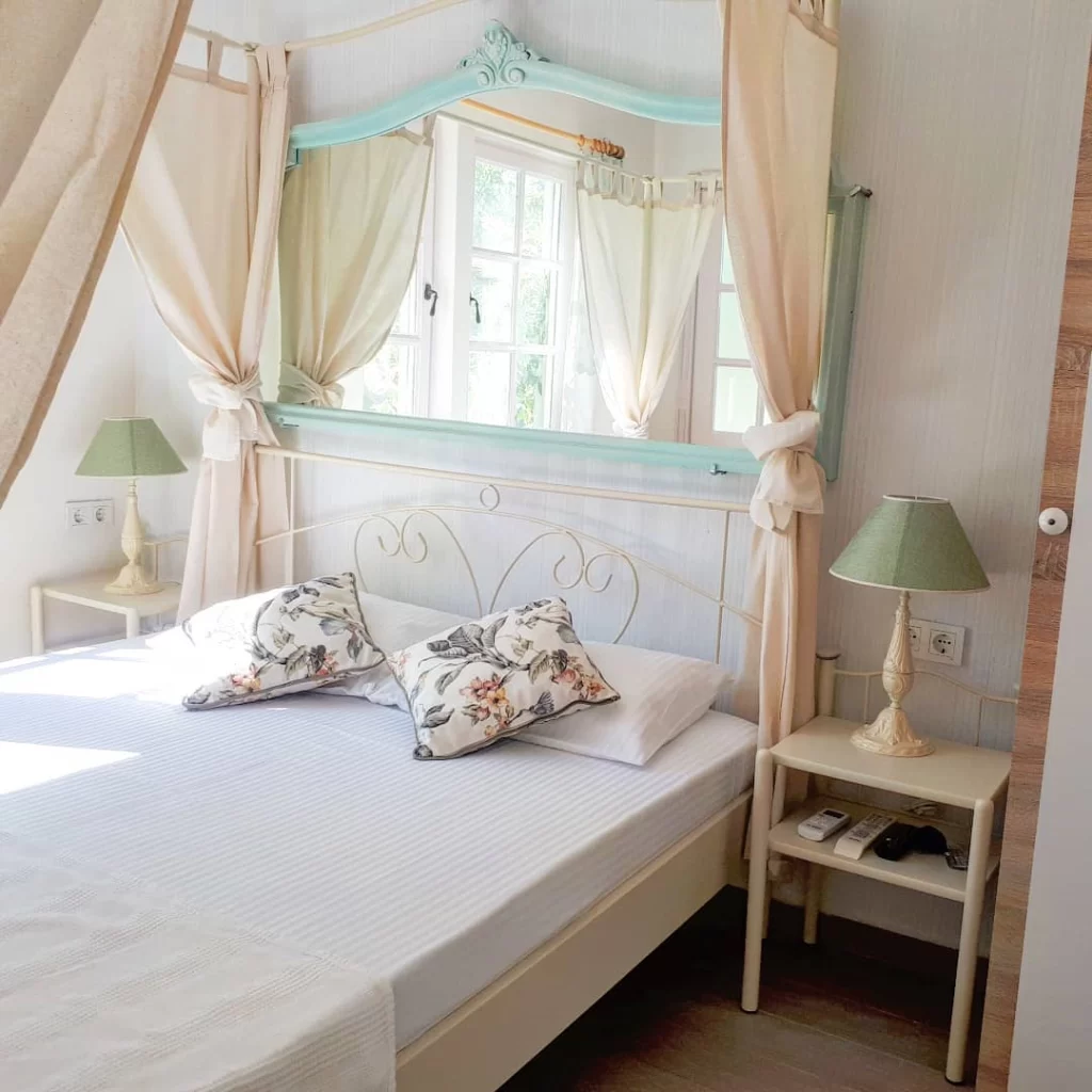 Beyaz ferforje başlıklı, üzerinde çiçek desenli yastıklar bulunan konforlu bir çift kişilik yatak, Bozcaada'nın romantik atmosferini yansıtan aydınlık bir otel odasında, aynadan yansıyan yeşil ağaçlarla huzurlu bir kaçış rotası sunuyor.