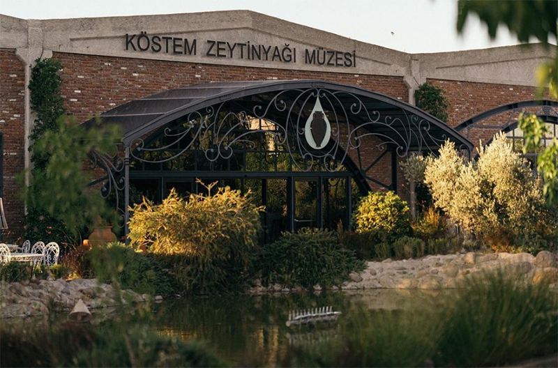 Urla'nın bereketli doğası ve zeytincilik kültürüyle öne çıkan, önünde zeytin ağaçları ve yeşil bitkilerin bulunduğu bir göletin yer aldığı, tuğla duvarlı Köstem Zeytinyağı Müzesi'nin demir işlemeli giriş kapısı.