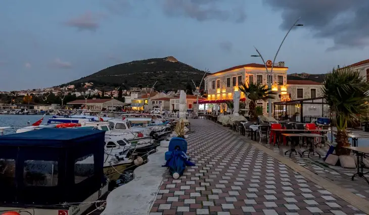 Alacakaranlıkta Ege'nin sevilen tatil rotası Foça'nın sahil şeridi, rıhtımda demirli tekneleri, ışıklı taş binaları ve açık hava masalarıyla dolu desenli bir gezinti yolu ile karşıdaki yeşil tepelerin oluşturduğu huzurlu manzarayı sergiliyor.