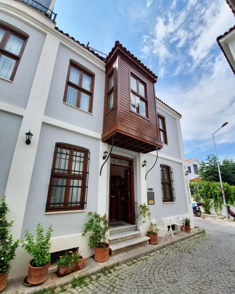 Bozcaada'nın Rum Mahallesi'nde yer alan, açık gri renkli duvarları ve ahşap cumbalı penceresiyle dikkat çeken Latife Hanım Konağı'nın geleneksel mimarisi, Arnavut kaldırımlı sokakta sıralanmış saksı bitkileri ve dışarıda duran beyaz masa sandalye ile huzurlu bir Bozcaada gezisi atmosferi sunuyor.