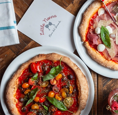Ahşap bir masa üzerinde, taze renkli çeri domatesler, siyah zeytinler ve fesleğen yapraklarıyla dolu glutensiz bir pizza ile dilimlenmiş jambon, küçük mozzarella topları ve rendelenmiş parmesan peyniri içeren bir başka pizza, Paris restoranları arasında özel bir yere sahip Little Nonna'nın lezzetlerini sergiliyor.