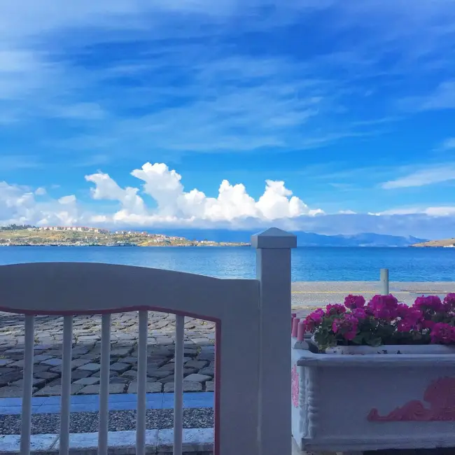 Masmavi Ege Denizi'nin berrak sularına nazır, beyaz süslü bir korkuluk ve canlı pembe çiçeklerle dolu saksılarla çevrili bu huzurlu Foça manzarası, karşı kıyıda uzanan şirin yerleşim yerleri ve gökyüzündeki pamuksu beyaz bulutlarla Ege'nin sevilen rotası Foça'nın davetkar atmosferini yansıtıyor.