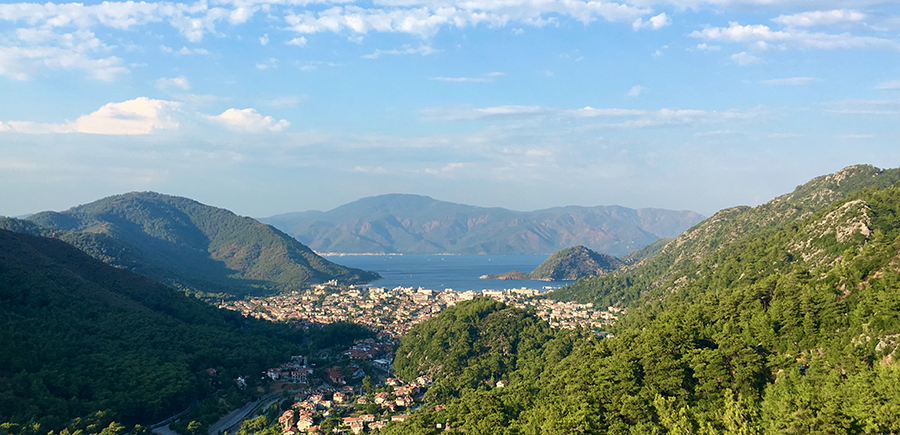 Yemyeşil çam ormanlarıyla çevrili tepeden görünen Marmaris'in yerleşim alanlarının masmavi körfez boyunca uzandığı, uzaktaki dağların ve açık gökyüzünün eşlik ettiği, bir Marmaris seyahat rehberi için ideal, büyüleyici panoramik bir manzara.