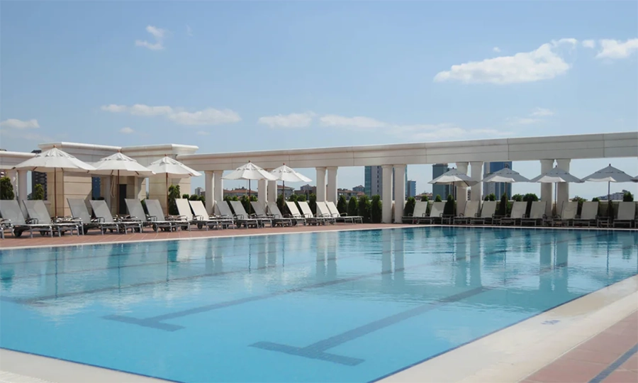 Ataşehir'deki Marriott Hotel Asia'nın, uzun mavi yüzme havuzunun bir tarafında beyaz şemsiyelerle gölgelenmiş çok sayıda boş şezlongun sıralandığı, arka planda şehrin yüksek binalarının göründüğü ve yazın İstanbul'da serin bir kaçamak sunan dinlendirici bir otel havuzu manzarası.