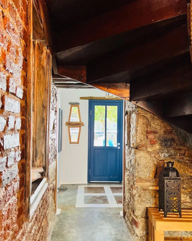 Kırmızı tuğla ve doğal taş duvarlarla çevrili, üzerinde koyu ahşap basamaklı merdivenlerin uzandığı, geleneksel ve modern detayları harmanlayan dar bir geçidin sonunda, dışarıya açılan cam panelli canlı koyu mavi bir kapı beliriyor; sol duvarda rustik ahşap çerçeveli dekoratif aynalar, sağda ise taş duvarın dibinde yer alan sade ahşap bir bank üzerinde oymalı metal bir fener duruyor, zemin ise ortasında açık renkli geometrik desenlerle süslenmiş olup, kapıdan süzülen doğal ışıkla sıcak ve davetkar bir giriş atmosferi yaratıyor.