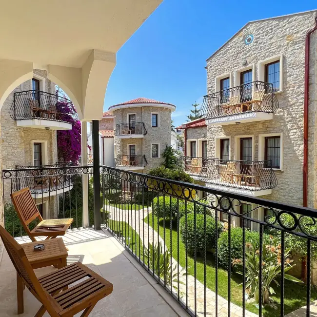 Ege ile Akdeniz'in kesişim noktasındaki Datça'da konaklama deneyimini yansıtan bu fotoğrafta, ahşap balkon sandalye ve masasının bulunduğu balkondan bakıldığında, taş dokulu otel binalarının yemyeşil bahçe ve canlı mor begonvillerle çevrili avlusu, masmavi bir gökyüzü altında huzurlu bir tatil ortamı sunuyor.
