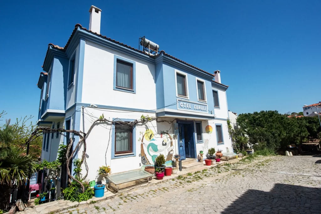 Mavi detaylarla süslü beyaz badanalı, iki katlı Otel Ebruli, Bozcaada'nın taş sokaklarından birinde, yöresel mimarisiyle dikkat çeken bir butik otel olarak karşımıza çıkıyor; girişindeki renkli saksılar, bitkiler ve deniz temalı bir duvar resmiyle adanın sıcaklığını ve huzurunu yansıtan bu aile işletmesi, romantik bir kaçış rotası arayan misafirlerine kapılarını açıyor.