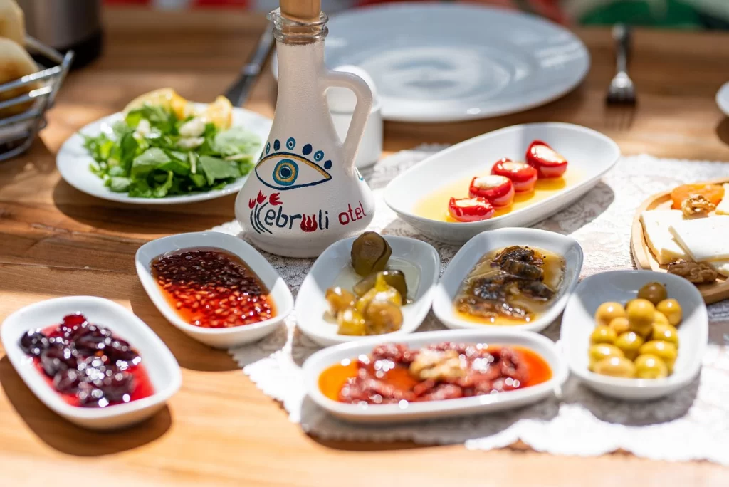 Bozcaada'nın Kuzey Ege'deki romantik kaçış rotasında, Otel Ebruli'nin ahşap bir masa üzerinde, el yapımı reçeller, zeytinler, peynirler ve özel soslarla dolu rengarenk bir organik kahvaltı servisi; ortada nazar boncuklu Ebruli Otel yazılı sürahi ve arka planda taze yeşillikler ile iştah açıcı bir görüntü sunuyor.