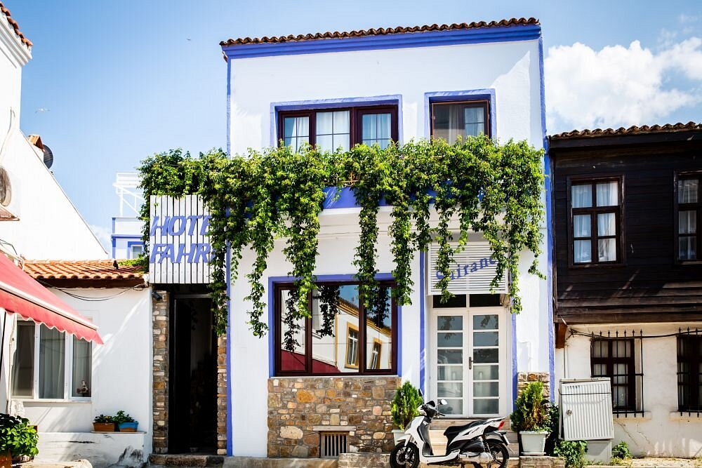 Bozcaada'nın karakteristik mimarisine sahip, mavi detayları ve sarkan yeşillikleriyle dikkat çeken Hotel Fahri'nin girişi, önünde park etmiş beyaz bir scooter ile bu Ege adasındaki romantik konaklama deneyimini vurguluyor.