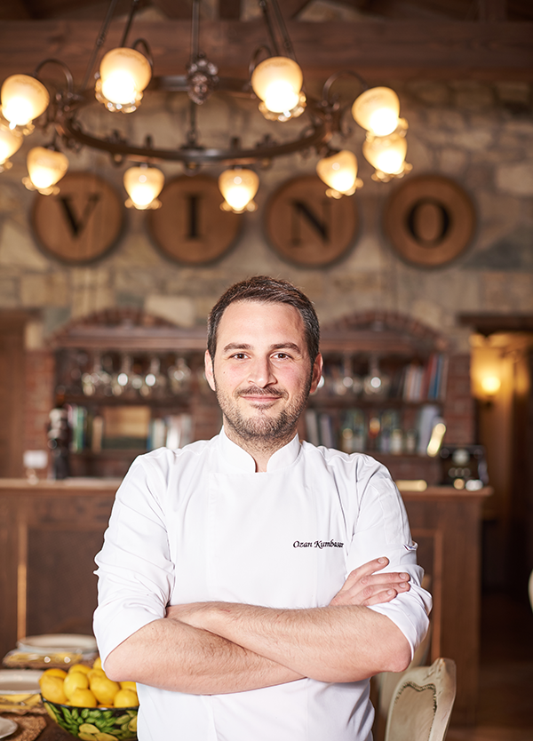 Urla'nın meşhur Bağ Rotaları üzerinde yer alan, doğal ve otantik atmosferiyle dikkat çeken Vino Locale restoranının şefi Ozan Kumbasar, beyaz aşçı ceketini giymiş, VINO yazılı ahşap varillerin ve şık avizenin önünde, masada duran parlak limonların eşlik ettiği sıcak bir ortamda kameraya gülümseyerek poz veriyor.