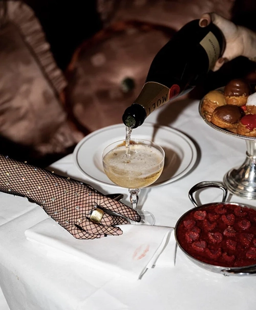 Şık bir Paris restoranında beyaz masa örtüsü üzerinde, parıltılı file eldivenli bir el ruj izi olan bembeyaz peçeteye zarifçe uzanırken, diğer bir el köpüklü şampanyayı klasik coupe bardağa dolduruyor; masada ayrıca rengarenk küçük pastalar ve taze ahududular dolu gümüş bir kâse bulunuyor.