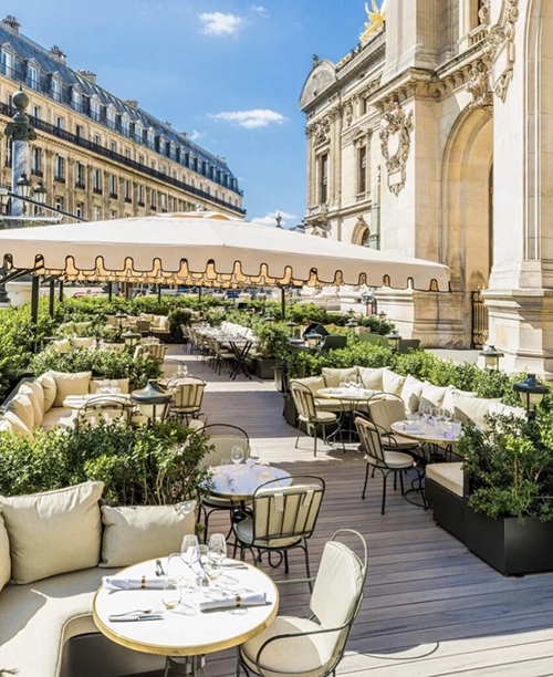 Krem rengi minderli oturma grupları, beyaz düzenlemeli masaları ve dantel kenarlı büyük tentesiyle şık bir açık hava Paris restoranı terası, saksılardaki gür yeşillikler ve arka planda görünen görkemli, süslü tarihi bir bina ile klasik Paris apartmanları arasında huzurlu ve lüks bir atmosfer sunuyor.