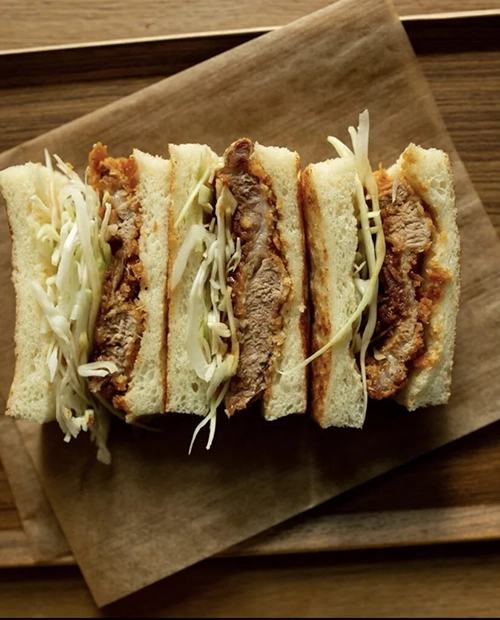 Ahşap bir zemin üzerindeki kahverengi parşömen kağıdında, çıtır pane et ve ince doğranmış lahana ile doldurulmuş beyaz tost ekmeğinden yapılmış, dört eşit parçaya bölünmüş bu sandviç, Paris restoranlarının dünya mutfaklarından sunduğu eşsiz lezzetlerden birine işaret ediyor.
