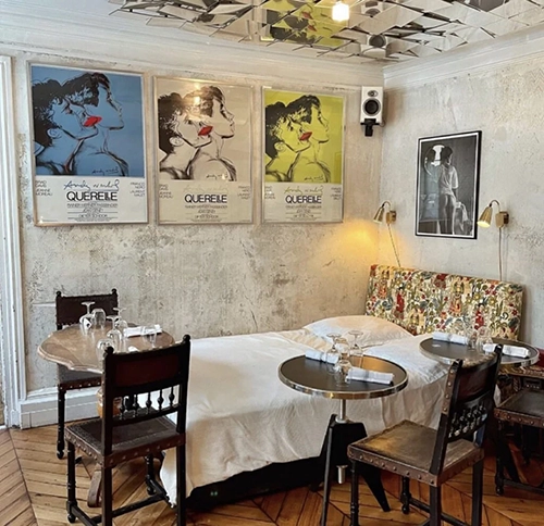 Bir yatağın etrafına yerleştirilmiş yemek masaları ve sandalyeleriyle sıra dışı bir Paris restoranı iç mekânı, pop art posterler, yatak başlığındaki desenli kumaş ve yansıtan ayna mozaik tavanıyla dikkat çekiyor.