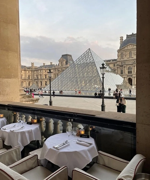 Paris'in kalbinde, Louvre Müzesi'nin ikonik cam piramidine nazır konumlanan şık bir restoranda, beyaz örtülü masalar ve konforlu koltuklarla donatılmış açık hava bölümünden, tarihi binalar ve hareketli meydanın keyifli manzarası sunulurken, bu huzurlu ambiyans En İyi Paris Restoranları deneyimlerinden birini yansıtıyor.