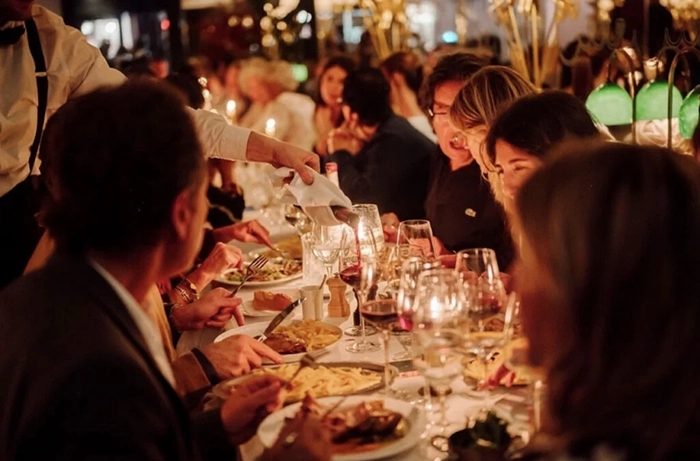 Şık bir Paris restoranında, loş ışıklı ve sıcak bir ortamda, beyaz masa örtülü uzun bir masanın etrafında toplanmış konuklar, bir garsonun şarap servisiyle zenginleşen bir akşam yemeği sırasında lezzetli yemeklerin ve keyifli sohbetin tadını çıkarırken, şehrin ünlü gastronomi deneyimini yansıtıyor.