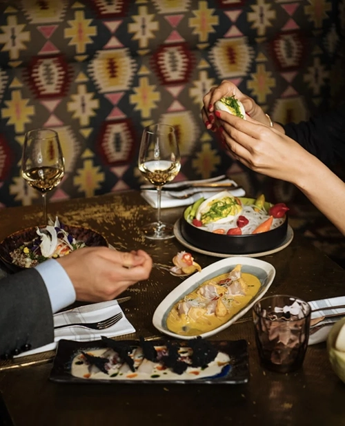 Geometrik desenli duvar önünde oturan iki kişinin, Paris restoranlarından birinde beyaz şarap eşliğinde ceviche benzeri deniz ürünleri ve buharlı ekmek (bao bun) gibi özenle hazırlanmış farklı lezzetleri deneyimleyerek keyifli bir yemek anı yaşadığı görülüyor.