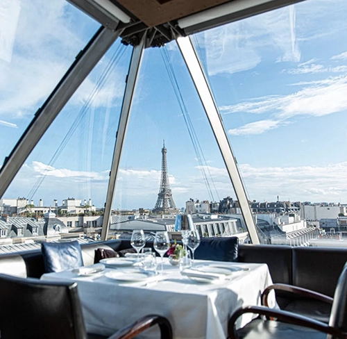 Modern cam tavanlı ve duvarlı bir Paris restoranının içinden, bembeyaz örtülü zarif masada şarap kadehleri ve tabaklar yer alırken, büyüleyici Eyfel Kulesi ve Paris'in ikonik çatıları açık mavi gökyüzü altında panoramik bir manzara sunuyor, bu da onu En İyi Paris Restoranları arasında eşsiz bir deneyim kılıyor.