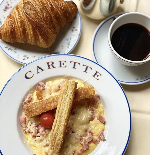 Carette restoranında servis edilen, üzerinde jambon ve erimiş peynir olan çırpılmış yumurtanın yanında çıtır bir kruvasan ve koyu demlenmiş kahveyle sunulan bu görsel, Paris'in en iyi kahvaltı deneyimlerinden birini gözler önüne seriyor.