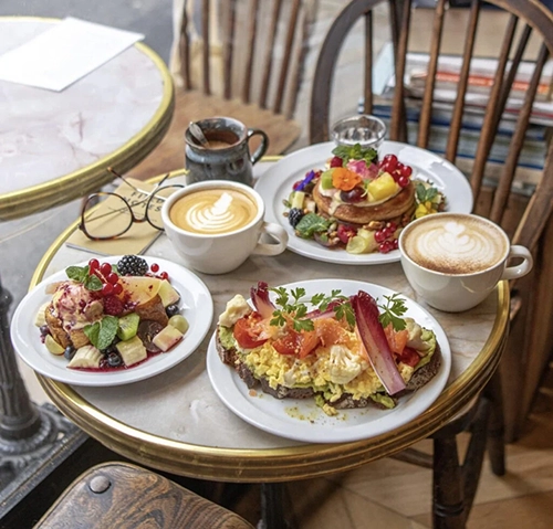 Mermer görünümlü bir masanın üzerinde, latte artlı kahveler, taze meyvelerle süslenmiş pankekler ve avokadolu omlet tost gibi özenle hazırlanmış çeşitli kahvaltılıkların sunulduğu bu masa, Paris'in en iyi restoranlarından birinde keyifli bir brunch deneyimini yansıtıyor.