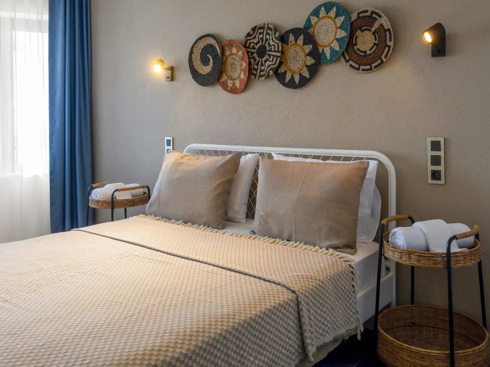 Bozcaada'daki romantik kaçış rotası konseptine uygun olarak tasarlanmış bu otel odasında, bej renkli yatak takımları ve beyaz metal karyolalı çift kişilik yatağın iki yanında rulo havlularla rattan komodinler, duvarda renkli hasır duvar süsleri ve pencerede mavi perdeyle sıcak, davetkar bir atmosfer sunuluyor.