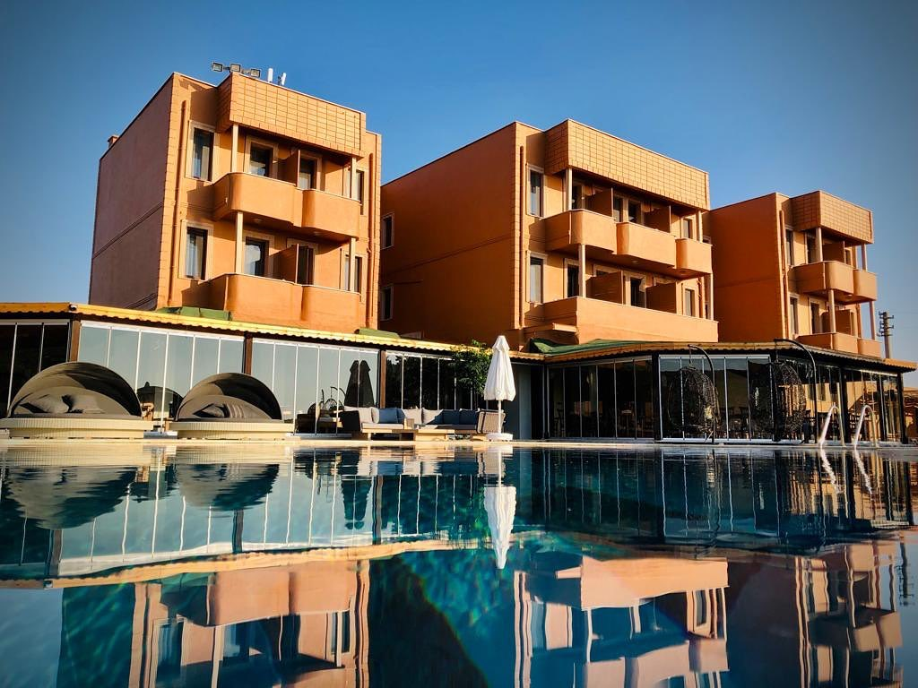 Bozcaada'nın huzurlu atmosferinde, berrak mavi gökyüzü altında parlayan Pelagos Hotel'in modern tasarımlı, şeftali renkli katlı binası ve havuz başında dinlenme alanlarının su yüzeyine yansıyan büyüleyici görüntüsü, burayı ideal bir kaçış rotası yapıyor.