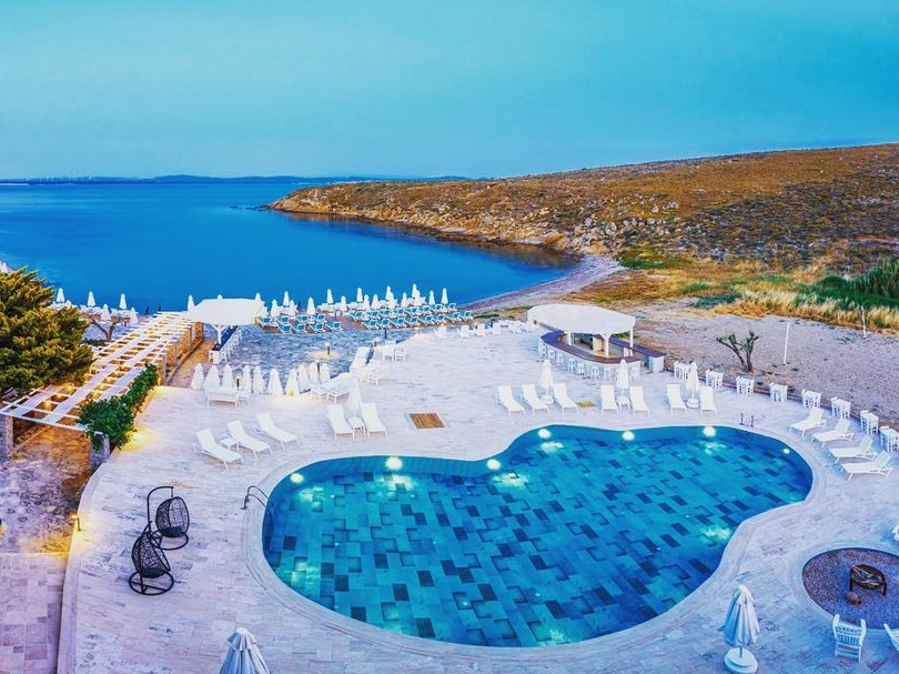 Bozcaada'nın kendine ait koyunda konumlanan Pelagos Otel'in havuz ve şezlonglarla dolu geniş terasından, Ege Denizi'nin berrak mavi koyuna açılan özel plajının ve onu çevreleyen doğal tepelerin huzurlu ve romantik kaçış rotası sunan manzarası.