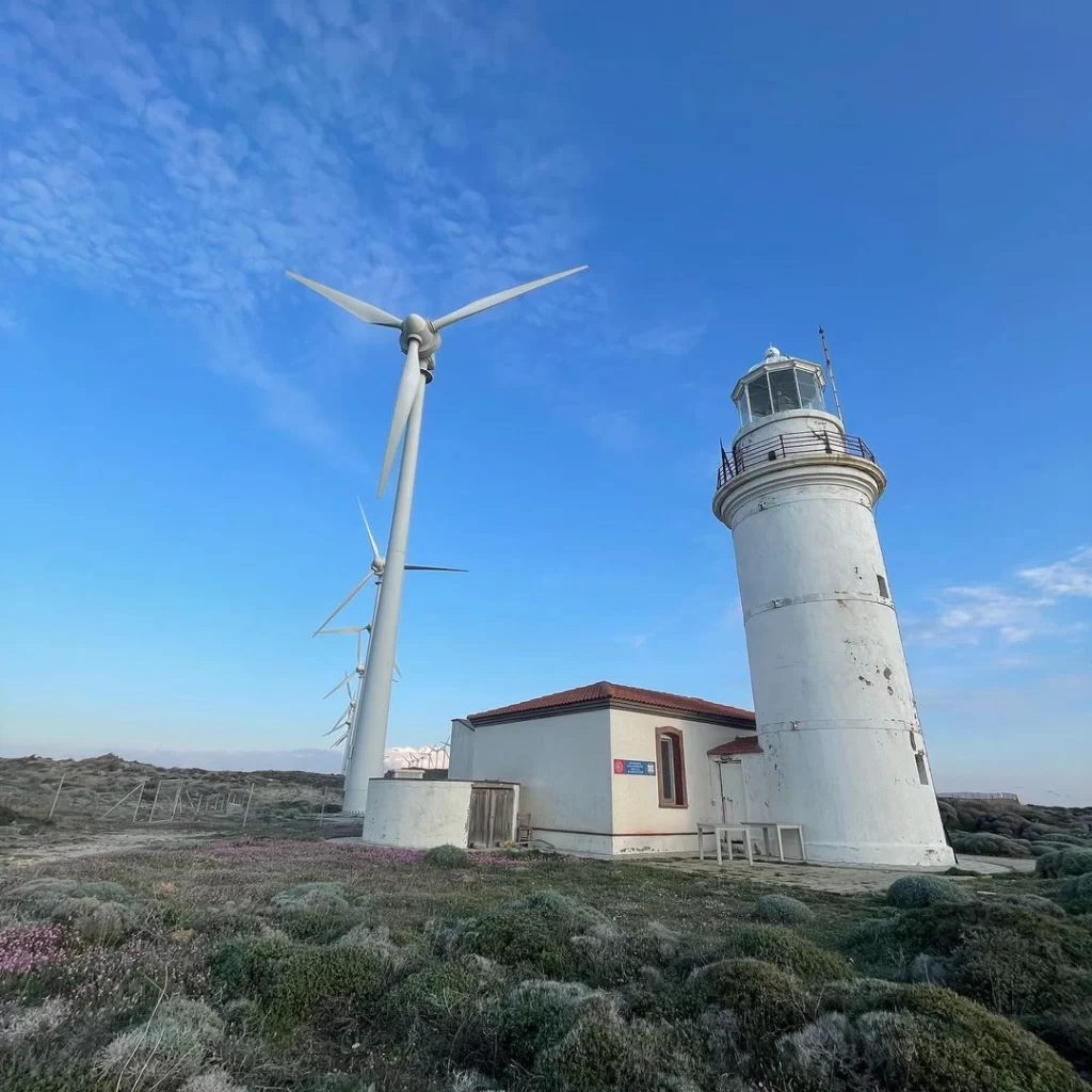 Kuzey Ege'nin romantik kaçış rotası Bozcaada'nın batı ucunda, yemyeşil bodur bitki örtüsüyle çevrili alanda, hafif yıpranmış beyaz Polente Feneri ve enerji üreten modern rüzgar gülleri, masmavi gökyüzü altında adanın ikonik gün batımı manzaralarını simgeleyen huzurlu bir tablo oluşturuyor.