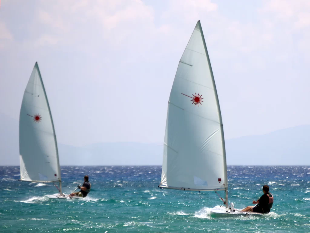 Kuzey Ege'nin Bozcaada açıklarında, rüzgarlı ve köpüklü turkuaz sularda iki sporcunun yelkenlileriyle hızla ilerleyerek adanın su sporları için sunduğu ideal koşulları gözler önüne serdiği dinamik bir manzara.
