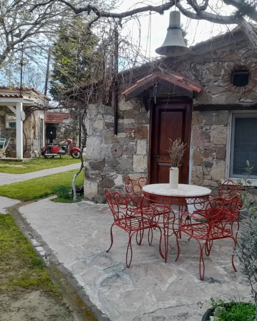 Taş duvarlı ve ahşap kapılı eski bir binanın önünde, kırmızı ferforje sandalyelerin çevrelediği yuvarlak bir masanın üzerinde kuru lavantalar ve tepesinde asılı çan, Bozcaada'nın huzurlu kaçış rotası atmosferini yansıtan yemyeşil bahçeli bir butik otelin dış mekanını gözler önüne seriyor.