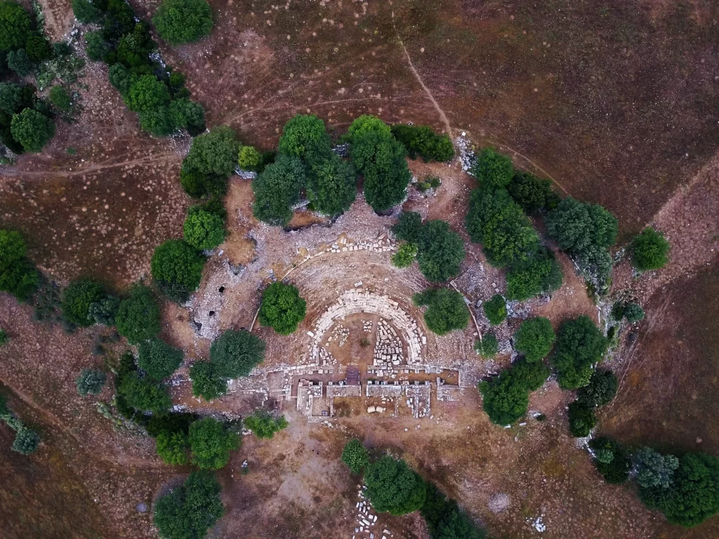 Kuşbakışı görünümdeki Teos Antik Kenti'nin kalıntıları arasında, yarı dairesel formda sıralanmış taş oturma yerleri ve sahne yapılarıyla eski bir tiyatro veya odeon, kurumuş toprak ve aralara serpişmiş yeşil ağaçlarla çevrili bir alanda belirginleşiyor.