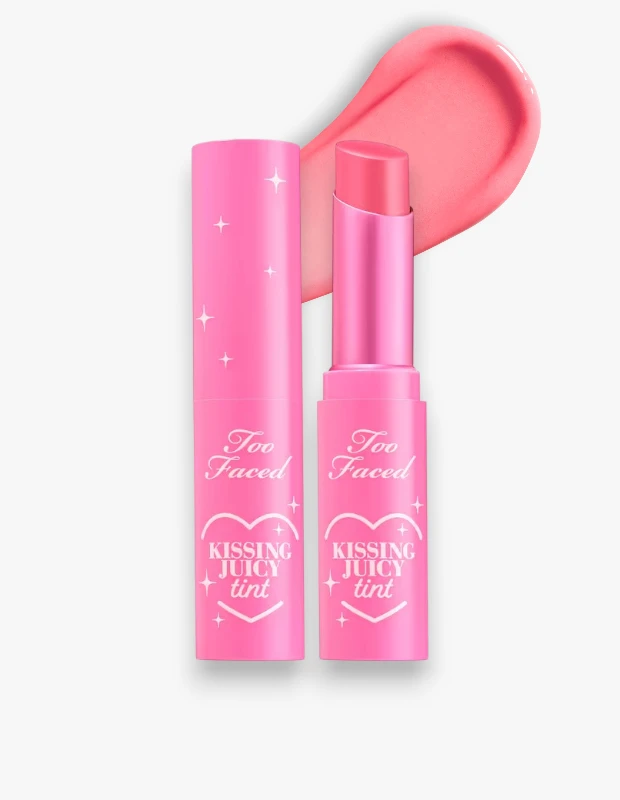 Beyaz bir arka plan üzerinde duran, Too Faced Kissing Juicy Tint adlı dudak ürününün ışıltılı pembe ambalajlı kapalı bir tüpü ve açık, hafif açılı ucuyla belirginleşen parlak şeker pembesi tonundaki çubuğu, arkasında ise ürünün nemli ve canlı dokusunu vurgulayan belirgin bir renk örneği sergilenmektedir.