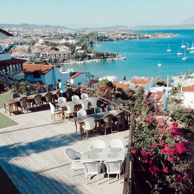 Datça'da, Ege'nin masmavi sularına nazır bir tepede konumlanan, zarif masa ve sandalyelerle döşenmiş geniş restoran terası, yemyeşil begonvillerin çevrelediği, denizde yüzen yelkenlilerle dolu Datça Körfezi ve sahildeki beyaz binaların oluşturduğu büyüleyici manzarayı gözler önüne seriyor.