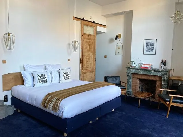 Bordeaux'daki tarihî Casa Blanca B&B'nin orijinal dokusunu koruyarak yenilenmiş odalarından biri: koyu mavi bazalı konforlu yatak, üzerinde dekoratif yastıklar ve sarı örtüyle davetkar bir atmosfer sunarken, odanın sağında yeşil mermer şömine, iki koltuk ve sanat eserleri, solunda ise rustik ahşap sürgülü kapı bu özgün balayı rotası deneyimini tamamlıyor; en güzel balayı rotaları için ilham veren bir kare.