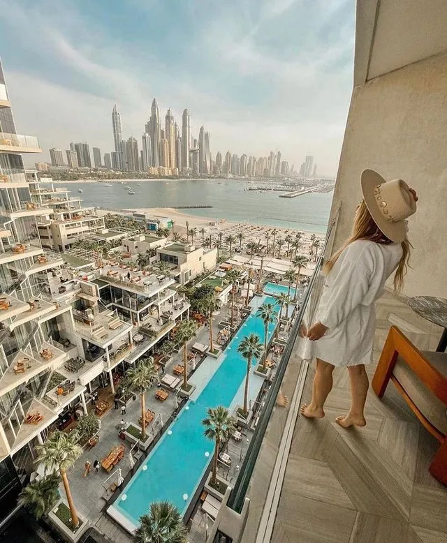 Yüksek bir balkonda beyaz bornoz ve şapka giymiş bir kişi, palmiye ağaçlarıyla bezeli uzun bir yüzme havuzuna, kumlu plaja ve arkadaki etkileyici Dubai şehir silüetine bakarak, lüks bir balayı rotası için ilham verici bir manzarayı seyrediyor; en güzel balayı rotaları için ilham veren bir kare.