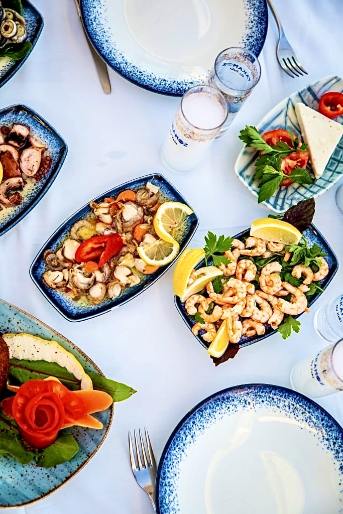 Bozcaada'nın beyaz masa örtülü sofrasında, Yakamoz markalı rakı kadehleri eşliğinde, bol limonlu karides, ahtapot salatası, deniz tarağı ve peynir gibi Ege mutfağına özgü taze deniz ürünleri ve meze çeşitlerinin mavi benekli tabaklarda iştah açıcı bir şekilde sunulduğu bu görsel, adanın zengin gastronomik deneyimini ve romantik kaçış rotası atmosferini gözler önüne seriyor.