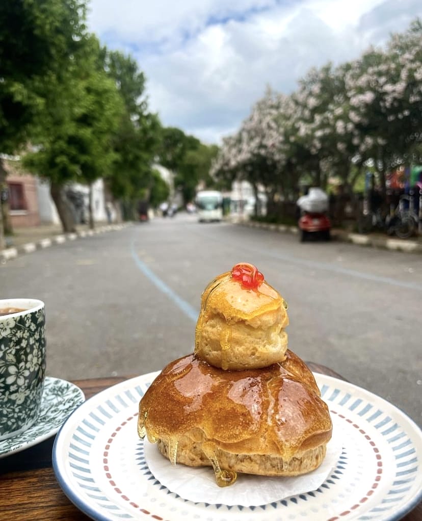 Desenli bir tabağın üzerinde, bal benzeri şerbetle bolca sırlanmış, altında büyük bir çörek, üstünde kırmızı bir vişne ile süslenmiş daha küçük bir çörek bulunan, iştah açıcı bir tatlı; solunda desenli bir fincan ile arka plandaki bulanık ağaçlı, araçlı sokak manzarası eşliğinde görünüyor.