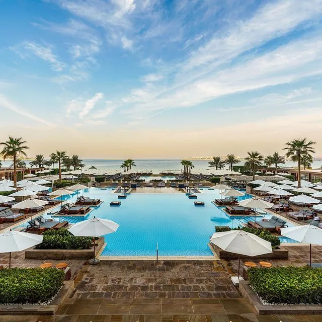 Dubai'deki Rixos Premium'un palmiye ağaçları ve beyaz şemsiyelerle süslenmiş, denize nazır geniş ve lüks havuz alanı, uzaktaki şehir silüetine karşı berrak turkuaz sularıyla romantik bir balayı rotası olarak çiftlere sakin ve unutulmaz bir deneyim sunuyor; en güzel balayı rotaları için ilham veren bir kare.