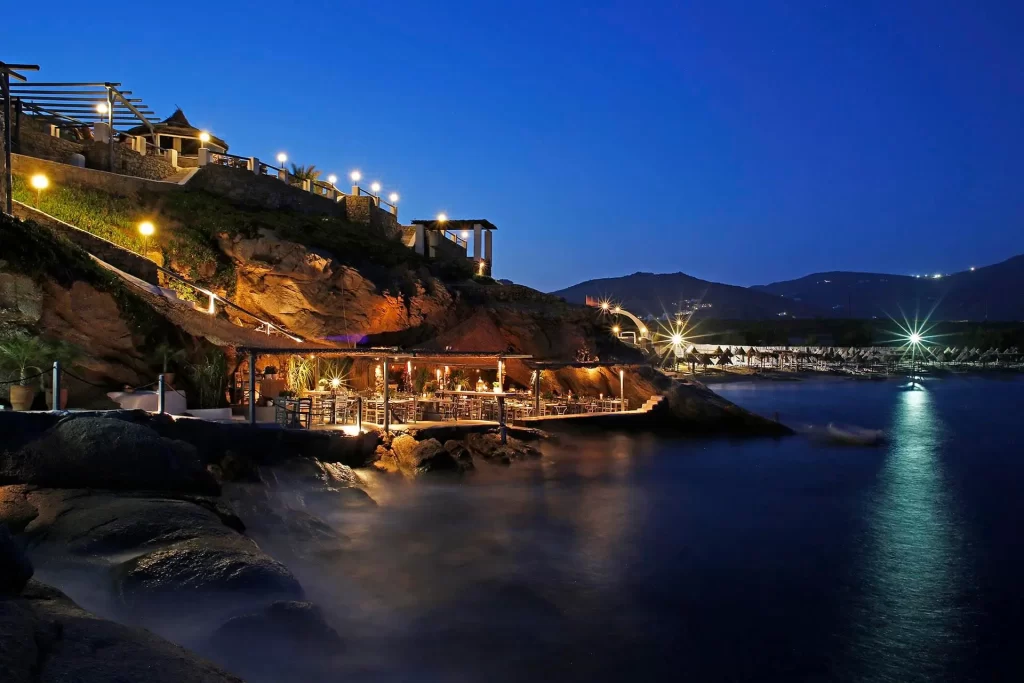 Mikonos'un hareketli Ege atmosferinde, kayalık bir yamaca kurulmuş, akşam ışıklarıyla parıldayan Spilia restoranı ve denize yansıyan ışıltıları, uzaklardaki sahilin ışıklarıyla birlikte büyüleyici bir gece manzarası sunuyor.