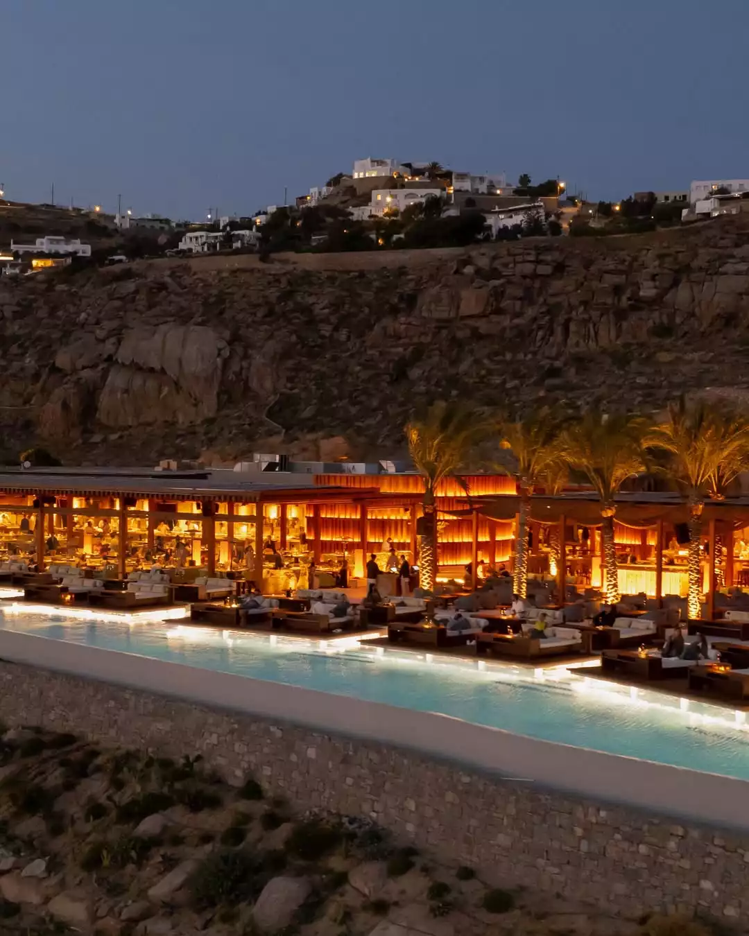 Gece çökerken, ışıl ışıl aydınlatılmış açık hava lüks restoran ve eğlence mekanının sonsuzluk havuzunun kenarında, şık misafirler sosyalleşirken, arka planda kayalık yamaçlara serpiştirilmiş yerleşim yerlerinin ışıkları beliriyor.