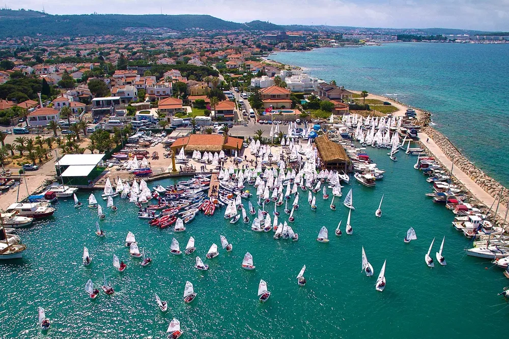Arkas Çeşme Yelken Kulübü'nün kalabalık bir etkinliğe ev sahipliği yaptığı bu havadan çekilen geniş açılı fotoğrafta, turkuaz renkteki denizde çok sayıda optimist sınıfı yelkenli tekne ve kıyıda sıralanmış binalar, palmiye ağaçları ve yeşil tepelerle çevrili hareketli bir liman manzarası görülüyor.
